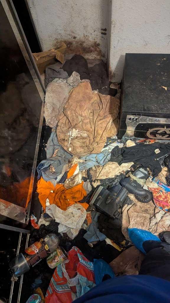 A cluttered indoor space with piles of clothing, plastic bottles, a vacuum hose and debris covering the floor. The wall behind is stained, and the area appears heavily neglected and unsanitary.