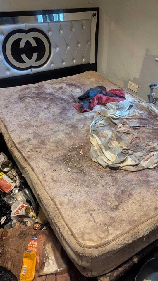 A dirty, cluttered bedroom with a stained mattress, crumpled bedding and rubbish scattered across the floor. Clothing, cans and debris are piled around the room, with a marked wall and an electrical outlet visible behind the bed.