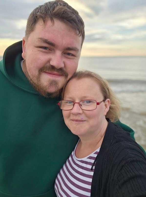 Oliver Turiccki and Sarah Pearce standing together outdoors near a body of water, both facing the camera. Turiccki wears a dark green hoodie; Pearce wears red glasses, a black cardigan and a striped top. The background shows calm water and a cloudy sky.