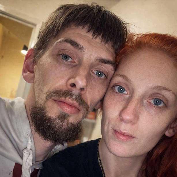 A close-up indoor selfie of a man with short brown hair and a beard beside a woman with red hair, both looking directly at the camera.
