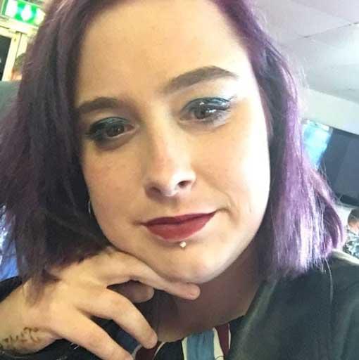A close‑up indoor portrait of Lisa Marie Riddell with short purple hair, red lipstick, dark eyeliner, and a piercing below her lower lip. She is resting her chin on her hand, with a green emergency exit sign visible in the background.