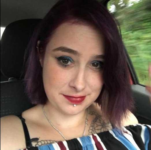 A portrait of Lisa Marie Riddell sitting in a car, wearing blue eyeshadow, red lipstick, and an off‑the‑shoulder striped top. She has short dark‑purple hair, a piercing below her lower lip, and tattoos on her upper chest and shoulder