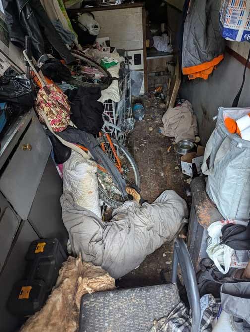 A cluttered, filthy interior space filled with debris, rubbish, and personal items scattered across the floor. The area is cramped, dirty, and chaotic, showing the squalid conditions in which two neglected dogs were kept.