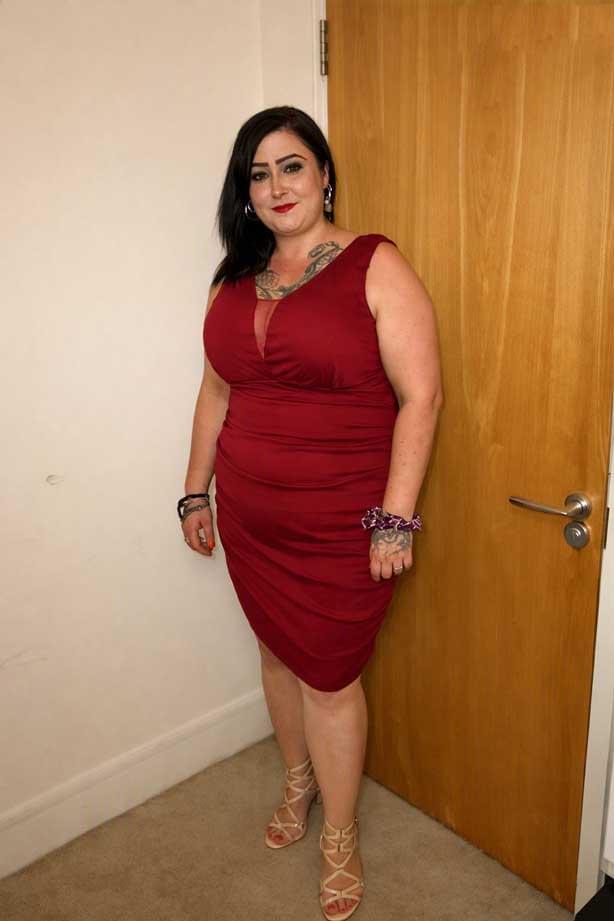 A full‑length photo of Lisa Marie Riddell standing near a wooden door, wearing a sleeveless burgundy dress and beige high‑heeled sandals. She has dark hair, visible tattoos on her chest and hand, and is wearing earrings and bracelets.