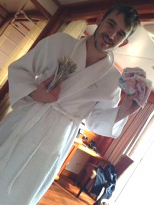Luke Wall sitting in a hotel room holding large wads of cash, posing for the camera with bundles of notes spread across his hands.