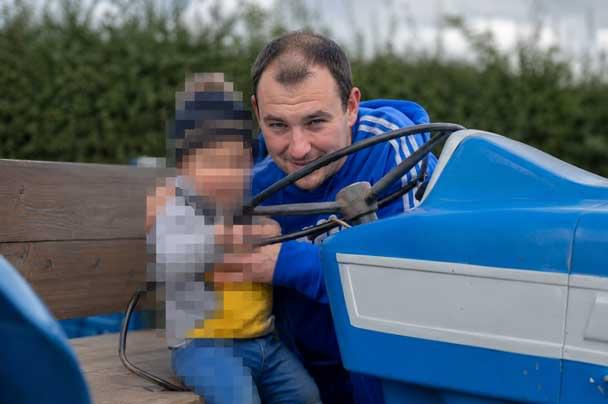 Matthew Shuker sits on a blue tractor outdoors, holding a small child whose face is pixelated. Greenery is visible in the background.