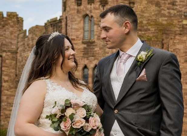 Matthew and Courtney Shuker pose together on their wedding day. Courtney wears a white lace gown and veil, holding a bouquet of roses, while Matthew wears a grey suit with a pink tie. They stand in front of a large stone building with arched windows.